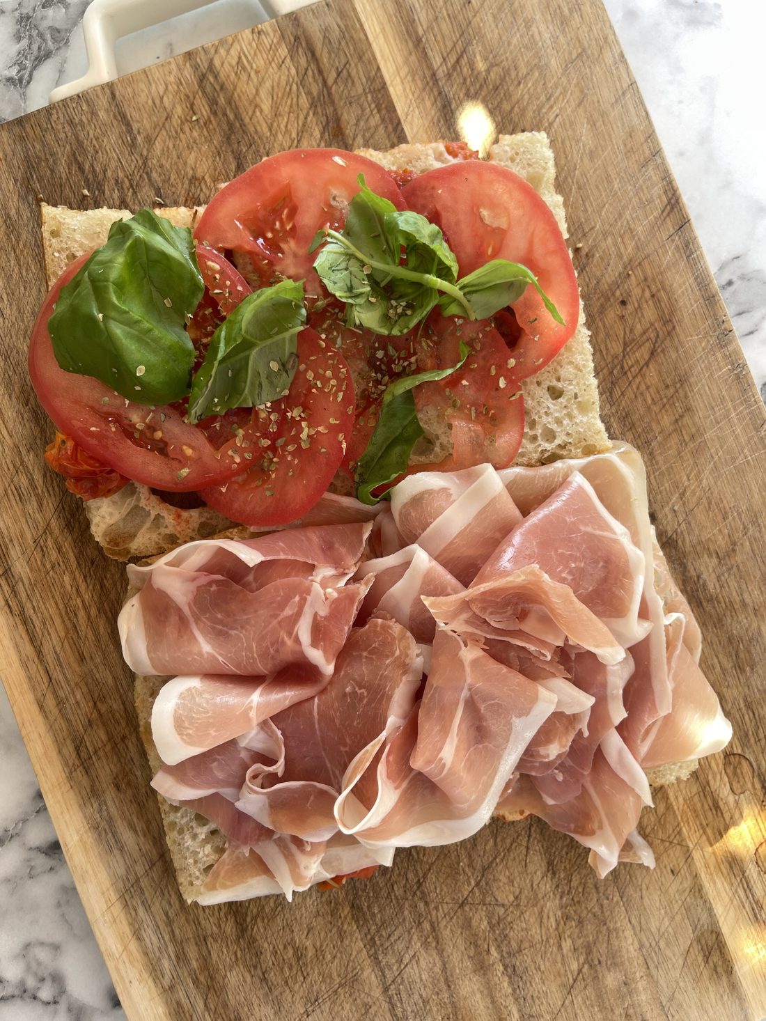 Przekrojona focaccia z plastrami pomidora, bazylią i szynką prosciutto na drewnianej desce