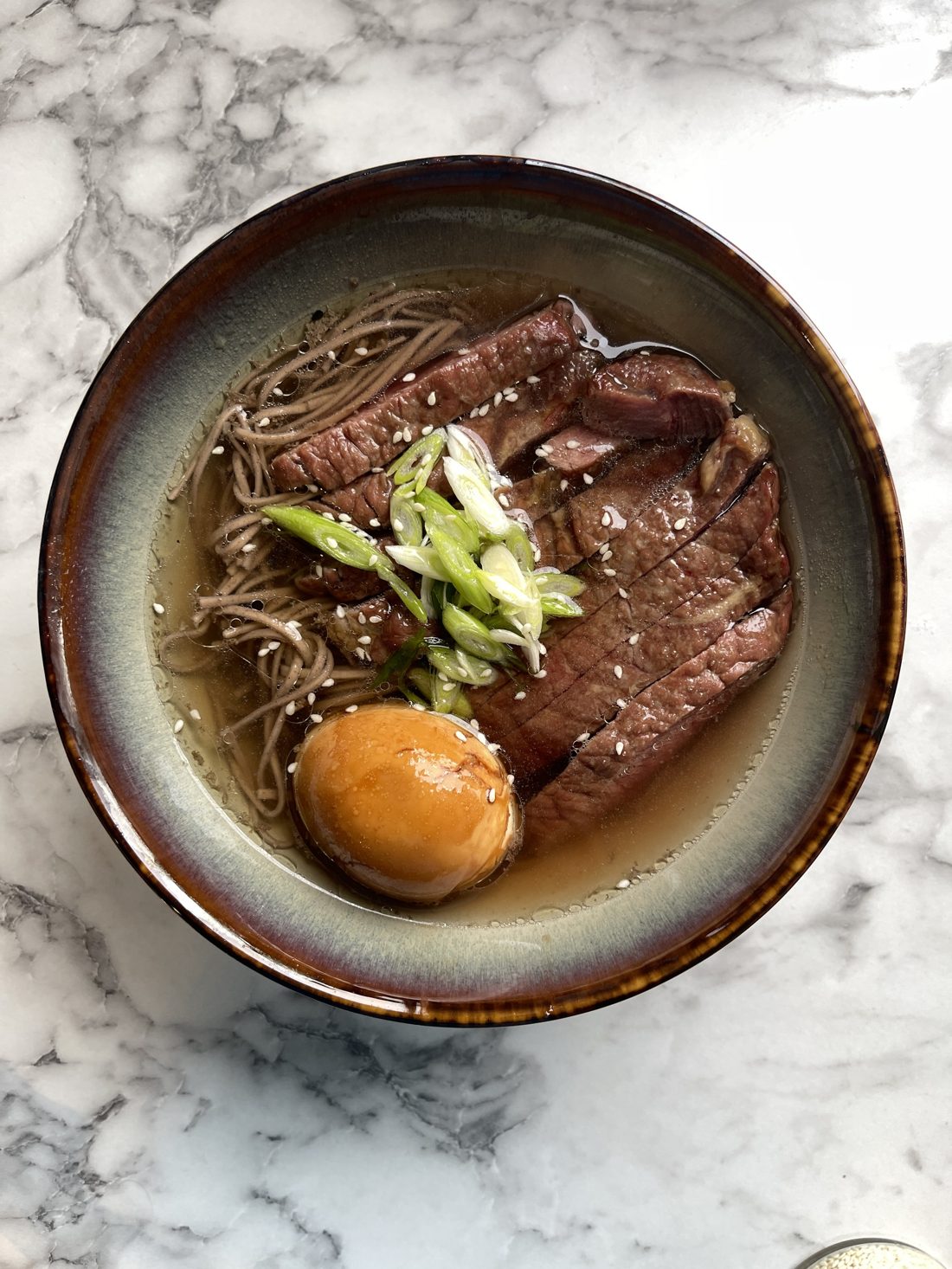 Miska makaronu soba w bulionie z plastrami wołowiny, marynowanym jajkiem, dymką i sezamem na marmurowym blacie