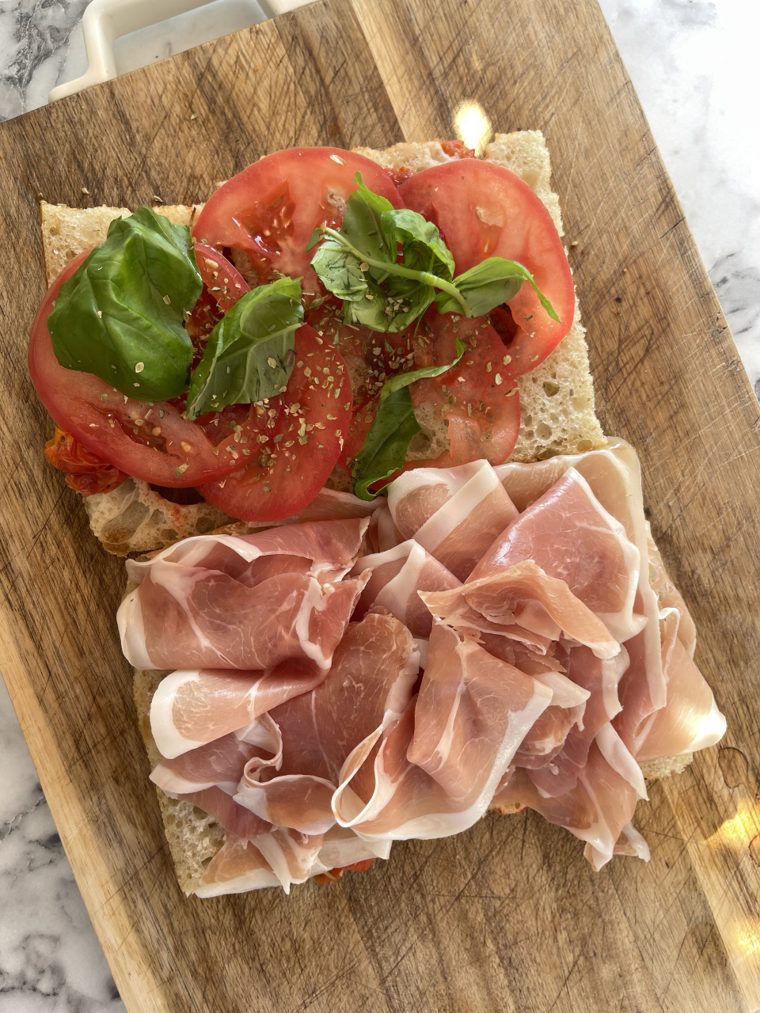 Przekrojona focaccia z plastrami pomidora, bazylią i szynką prosciutto na drewnianej desce
